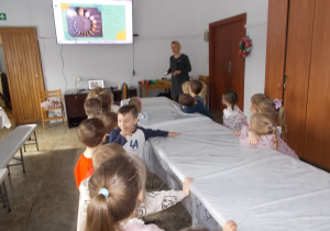 Dzieci siedzą przy długim stole. Przed nim stoi pani. Nad jej głową ekran z prezentacją wielkości różnych ptasich jaj.