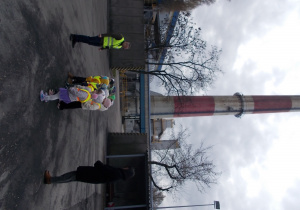 Dzieci stoją na dużym placu, w miejscu składowania węgla. Za nimi wysoki komin ciepłowni.