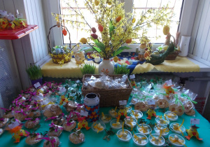 Stolik ustawiony przy oknie cudownie, wiosennie udekorowany: kwitnącą forsycją, zawieszonymi pisankami i ułożonymi pięknie zapakowanymi w folie i kolorowe wstążeczki produktami na kiermasz.