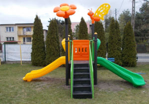 Nowa zjeżdżalnia. Z przodu wejście po metalowych schodkach, z obu boków część do zjazdów. Na górze przymocowane duże, plastikowe dekoracje: kwiatek i motylek.