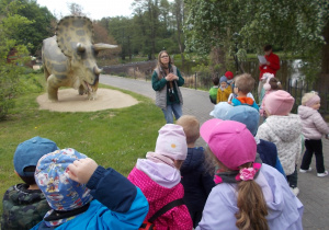 Dzieci stoją na ścieżce. Przed nimi pani przewodnik opowiada o umieszczonej za nią dużej figurze triceraptosa.