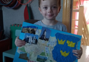 Chłopiec trzyma w dłoni obrazek a na nim duży herb miasta wypełniony plasteliną, dookoła przyklejone widokówki różnych zabytków.