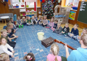 Dzieci siedzą w kole na dywanie. Na środku stoi białe waderko i stara walizka. Pan w bluzie gra na gitarze a dzieci śpiewają.