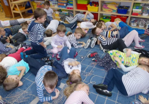 Dzieci leżą na dywanie i odpoczywają. Każde ma bluzkę w paski w różnych kolorach.