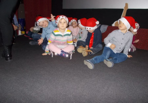 Grupka najmłodszych przedszkolaków siedzi na scenie. Na głowach mają mikołajkowe czapki.
