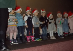 Te same dzieci stoją na scenie w szeregu. Niektóre mają otwarte buzie inne podnoszą ręce. Ilustrują ruchem piosenkę.