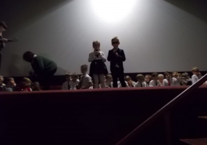 Duża scena. Z tyłu siedzą w szerego dzieci. Na środku dwoje konferansjerów. Z lewej strony dziewczynka trzymająca mikrofon, z prawej chłopiec w garniturze i ciemnych okularach.