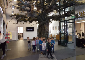 Duży hol. Na środku wysokie, metalowe drzewo. Pod nim grupka dzieci.