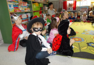 Uczestnicy balu siedzą na dywanie przy szafce z zabawkami. Na pierwszym planie dzieci w strojach: Zorro, biedroneczka, król.