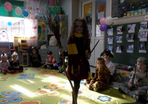Na środku dywanu stoi dziewczynka w stroju Hermiony z Harrego Pottera. W prawej ręce trzyma latającą miotłę w lewej różdżkę.