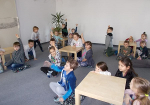 Na dywanie, przy małych stolikach siedzą dzieci. Niektóre podnoszą do góry rękę.