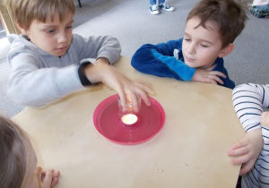 Na stoliku stoi talerz a na nim zapalona mała świeczka. Chłopiec przykrywa świeczkę szklanym naczyniem.