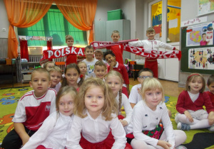 Dzieci siedzące na dywanie. Na drugim planie szaliki z napisem Polska.