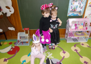 Dziewczynki w stroju myszki i kotka stoją i obejmują się. Dziewczynka w stroju zajączka siedzi przy nich na dywanie.
