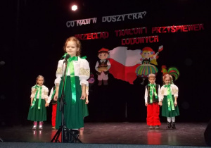 Na scenie. Dziewczynka stoi przy mikrofonie. Za nią, w dwóch trójkach stoją pozostałe dzieci.