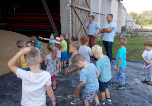 Dzieci stoją przed otwartymi drzwiami stodoły, z której wysypuje się zboże.