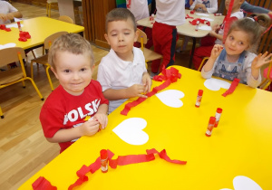 Przy stoliku dwóch chłopców i dziewczynka. Przed nimi leżą papierowe serduszka. W rękach trzymają paski czerwonej bibuły.