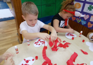 Chłopczyk i dziewczynka przy stoliku. Naklejają na swoje papierowe serduszka kulki wykonane z czerwonej bibuły.