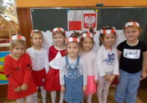 Wszystkie dziewczynki stoją przy tablicy. Uśmiechają się. Na głowach mają wianki w biało-czerwone kwiaty.