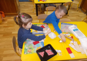 Chłopczyk i dziewczynka siedzą przy stoliku, malują połączone patyczki na czerwono, doklejają brodę z waty.