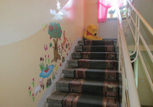 Klatka schodowa na piętro. Na ścianach kolorowe nalepki. Schody wyłożone chodnikiem.